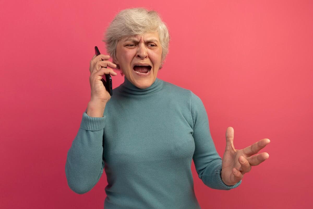 angry-old-woman-wearing-blue-turtleneck-sweater-talking-on-phone-keeping-hand-in-air-looking-down-1